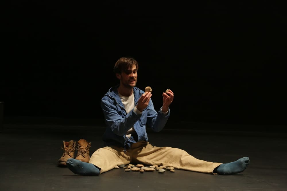 L'interprète est assis par terre jambes écartées. Il y a des cailloux posés entre ses jambes et il en tient un dans chaque main, à hauteur de son visage. Une paire de chaussures est posée à gauche à côté de lui