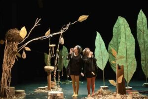 Dans un décor représentant des arbres avec des nids d'oiseaux, deux comédiennes soufflent dans des appeaux pour imiter le chant des oiseaux.