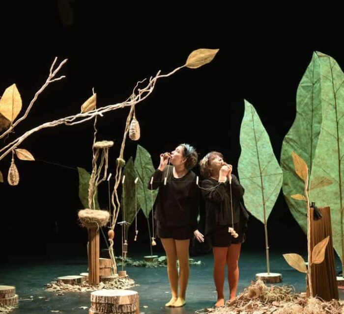 Dans un décor représentant des arbres avec des nids d'oiseaux, deux comédiennes soufflent dans des appeaux pour imiter le chant des oiseaux.