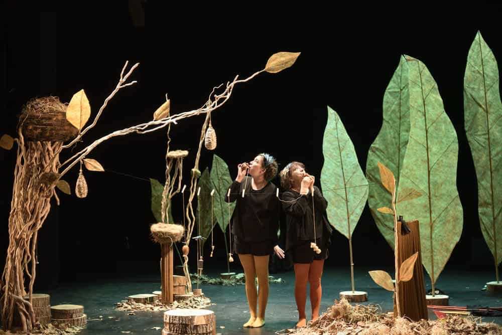 Dans un décor représentant des arbres avec des nids d'oiseaux, deux comédiennes soufflent dans des appeaux pour imiter le chant des oiseaux.