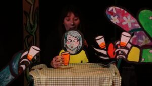 Une comédienne manipule une marionnette de papier représentant une petite fille à table avec un verre à la main.
