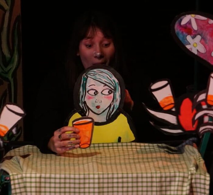 Une comédienne manipule une marionnette de papier représentant une petite fille à table avec un verre à la main.