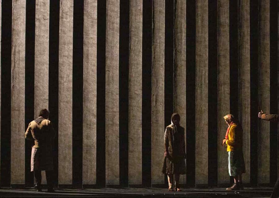 Le fond de la scène est rayé verticalement par des panneaux de tissus, la lumière est faible et arrive de la gauche de la scène. On distingue trois personnages en robes fleuries, gilets et foulards sur la tête. Un de dos, un de face et un de côté.