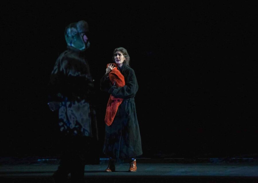 Une femme en manteau noir porte contre elle ce qui semble être un bébé emmailloté dans un châle rouge. Elle est tendue et regarde un homme en manteau noir et chapka.