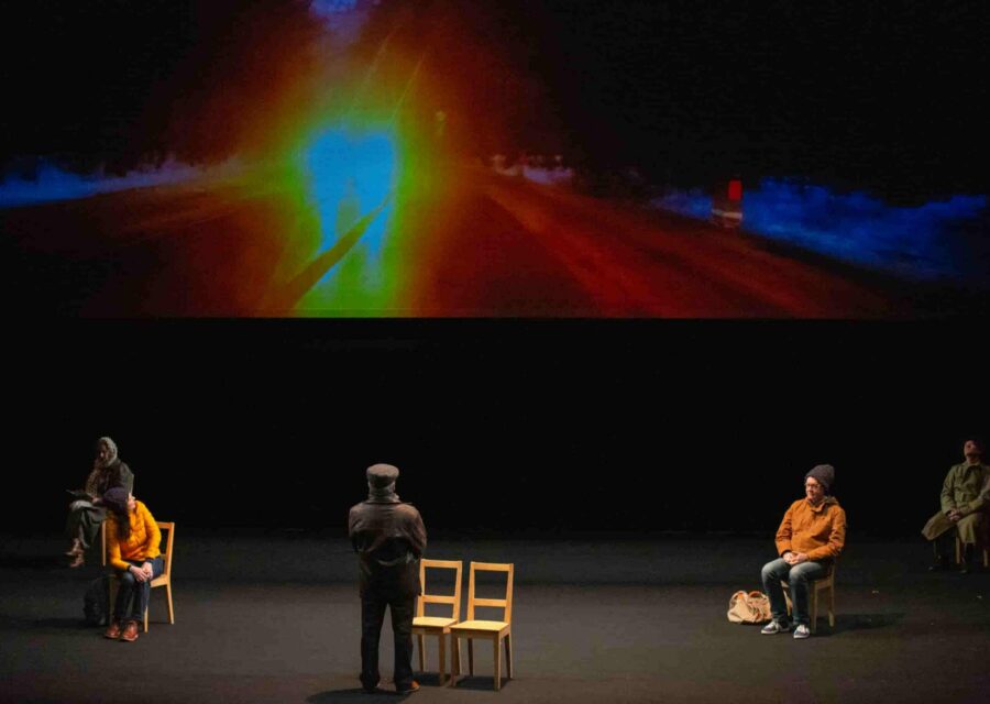 Un écran occupe le haut de la scène avec une route de nuit et les phares d’un véhicule. En dessous, deux comédiennes et deux comédiens sont assis sur des chaises, dos à l’écran. Une des comédiennes est en train de se retourner vers l’écran. Un cinquième comédien est debout au milieu devant deux chaises.