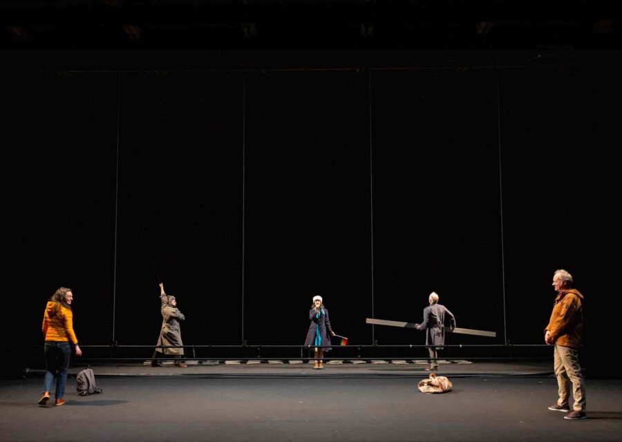 Cinq comédiens sont sur la scène vide à l’exception d’une perche descendue du gril technique. Au fond, on aperçoit des éléments qui vont composer le décor.