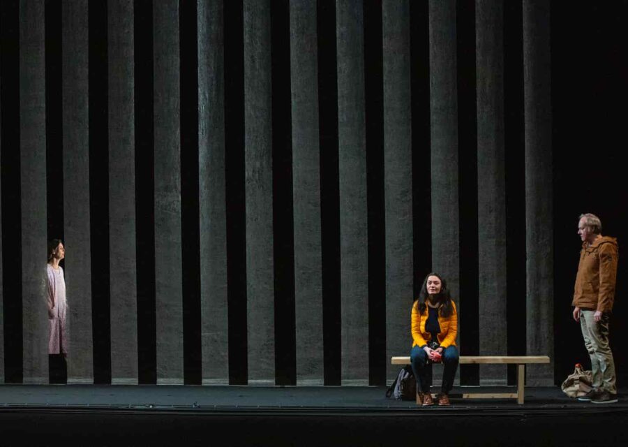 Le fond de la scène est rayé verticalement par des panneaux de tissus légèrement transparents. Une comédienne arrive de la gauche, derrière ces panneaux. Devant, à droite, une comédienne est assise sur un banc, face au public, un sac de voyage à ses pieds. Un comédien est debout à droite du banc avec un sac à ses pieds.