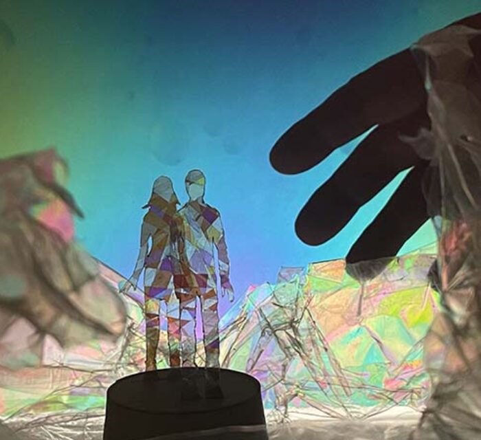 Deux silhouettes, un homme et une femme, posées sur un socle et entourées de matières colorés et transparentes. L'ombre d'une main géante apparaît à droite de l'image.