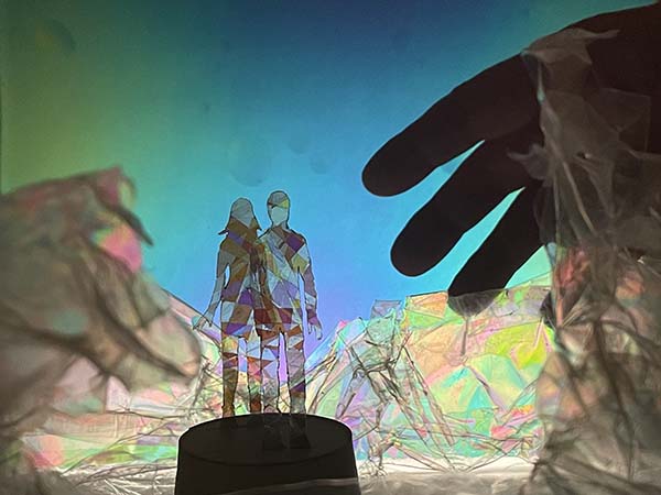 Deux silhouettes, un homme et une femme, posées sur un socle et entourées de matières colorés et transparentes. L'ombre d'une main géante apparaît à droite de l'image.
