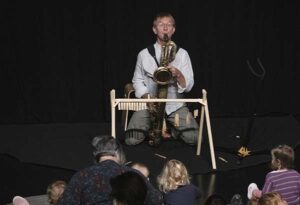 Des enfants et des adultes assis sur des coussins devant un musicien de face jouant du saxophone baryton. Il a devant lui un petit portique en bois sur lequel sont accrochés des anches et des bec de saxophone.