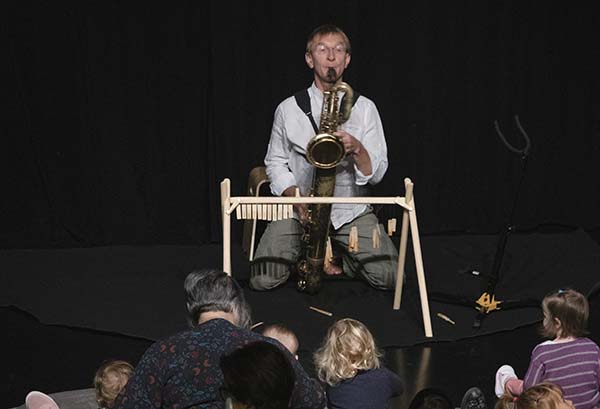 Des enfants et des adultes assis sur des coussins devant un musicien de face jouant du saxophone baryton. Il a devant lui un petit portique en bois sur lequel sont accrochés des anches et des bec de saxophone.