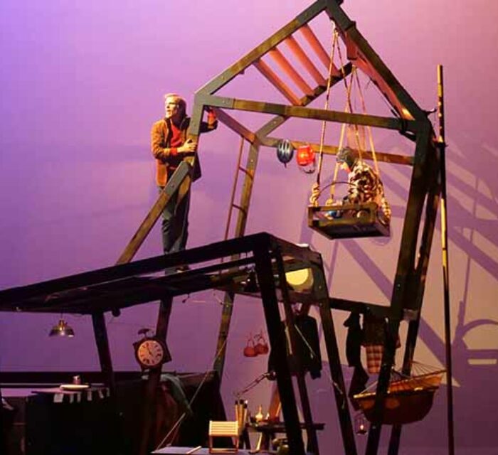Un décor de scène représentant l'ossature d'une maison bancale. Un homme est debout accroché à un des piliers. Un enfant est à droite assis sur une plateforme suspendue aux poutres du toit.