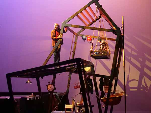 Un décor de scène représentant l'ossature d'une maison bancale. Un homme est debout accroché à un des piliers. Un enfant est à droite assis sur une plateforme suspendue aux poutres du toit.