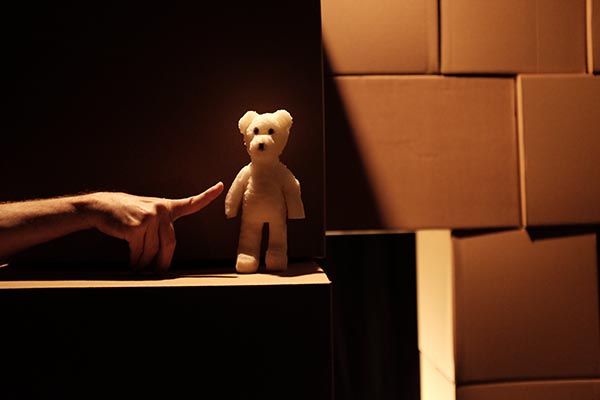 Une main pointe un doigt vers un ours en mousse devant un mur de cartons.