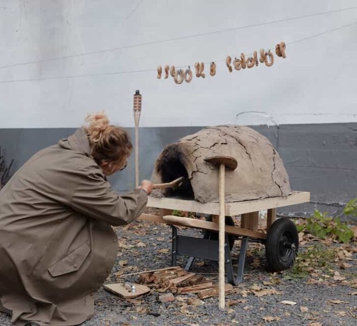 L'artiste fait cuire des petits pains dans un petits four en terre. Au-dessus d'elle, les petits-pains cuits sont suspendus sur un fil.