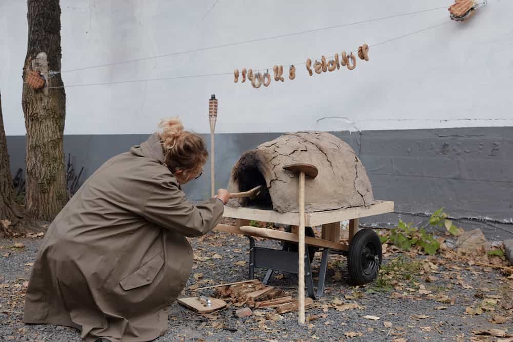 L'artiste fait cuire des petits pains dans un petits four en terre. Au-dessus d'elle, les petits-pains cuits sont suspendus sur un fil.