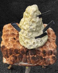 Un tabouret avec une assise en pain surmonté d'une énorme motte de beurre dans laquelle sont plantés 4 couteaux en forme de doigts.