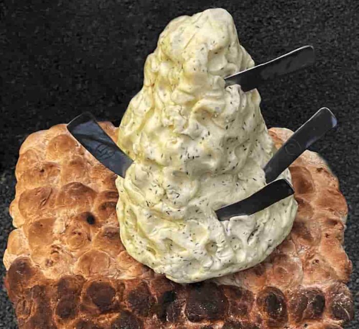 Un tabouret avec une assise en pain surmonté d'une énorme motte de beurre dans laquelle sont plantés 4 couteaux en forme de doigts.