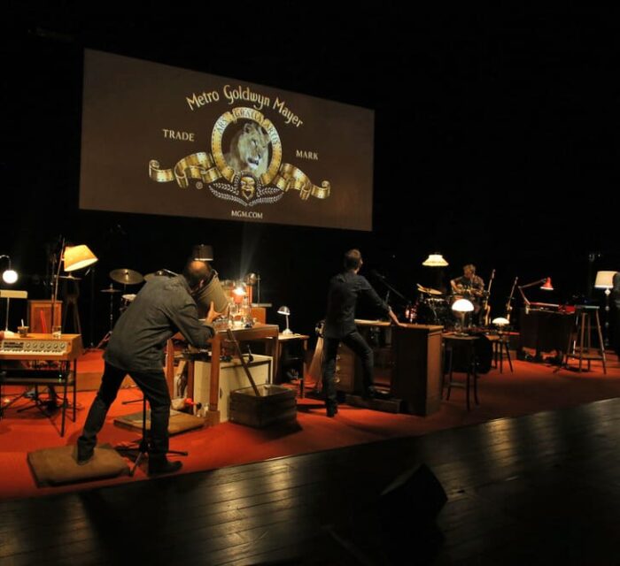 La scène est occupée par des instruments de musique et divers objets et un grand écran est installé en hauteur sur lequel on voit l'image du lion des génériques de la Métro Goldwyn Mayer. Les interprètes réalisent les bruitages et la musique en direct.