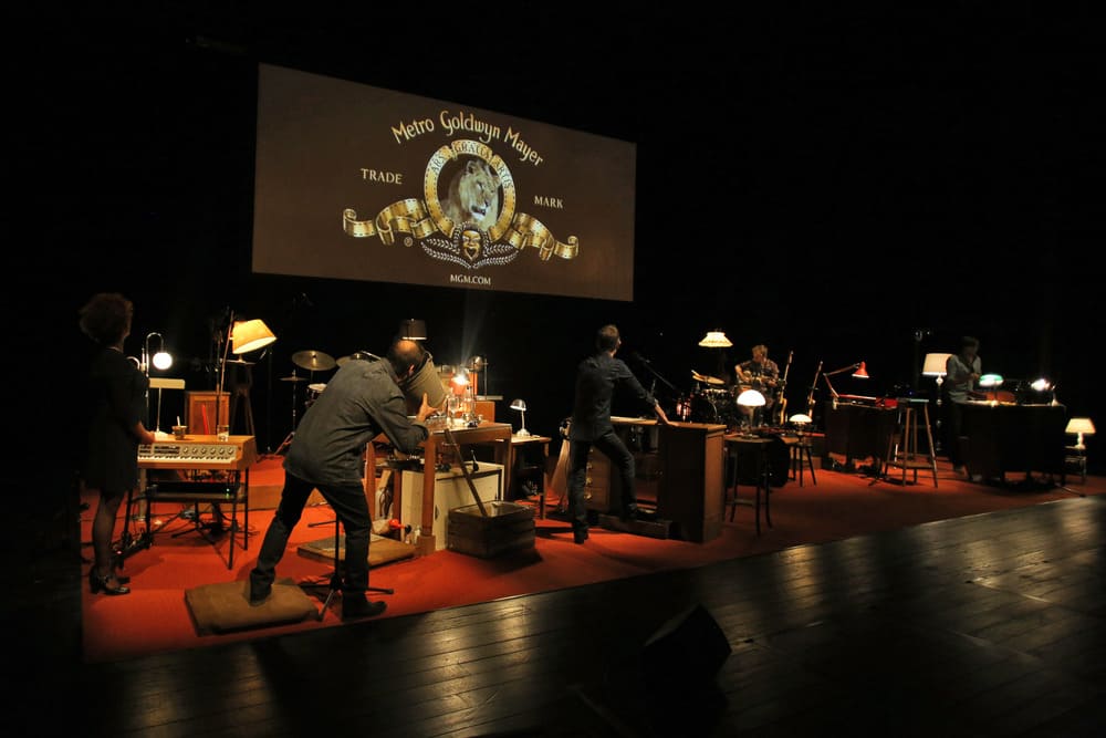 La scène est occupée par des instruments de musique et divers objets et un grand écran est installé en hauteur sur lequel on voit l'image du lion des génériques de la Métro Goldwyn Mayer. Les interprètes réalisent les bruitages et la musique en direct.