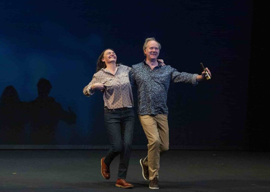 Une comédienne et un comédien marche sur la scène bras dessus, bras dessous. Le comédien tient une bière dans sa main gauche.