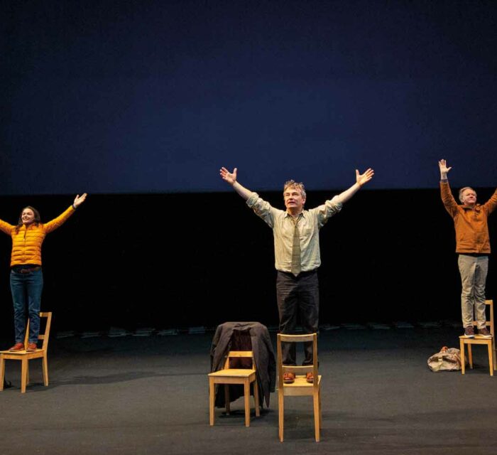 Une comédienne et deux comédiens sont chacun debout sur une chaise avec les bras en l'air.
