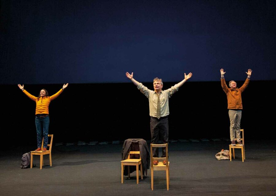 Une comédienne et deux comédiens sont chacun debout sur une chaise avec les bras en l'air.