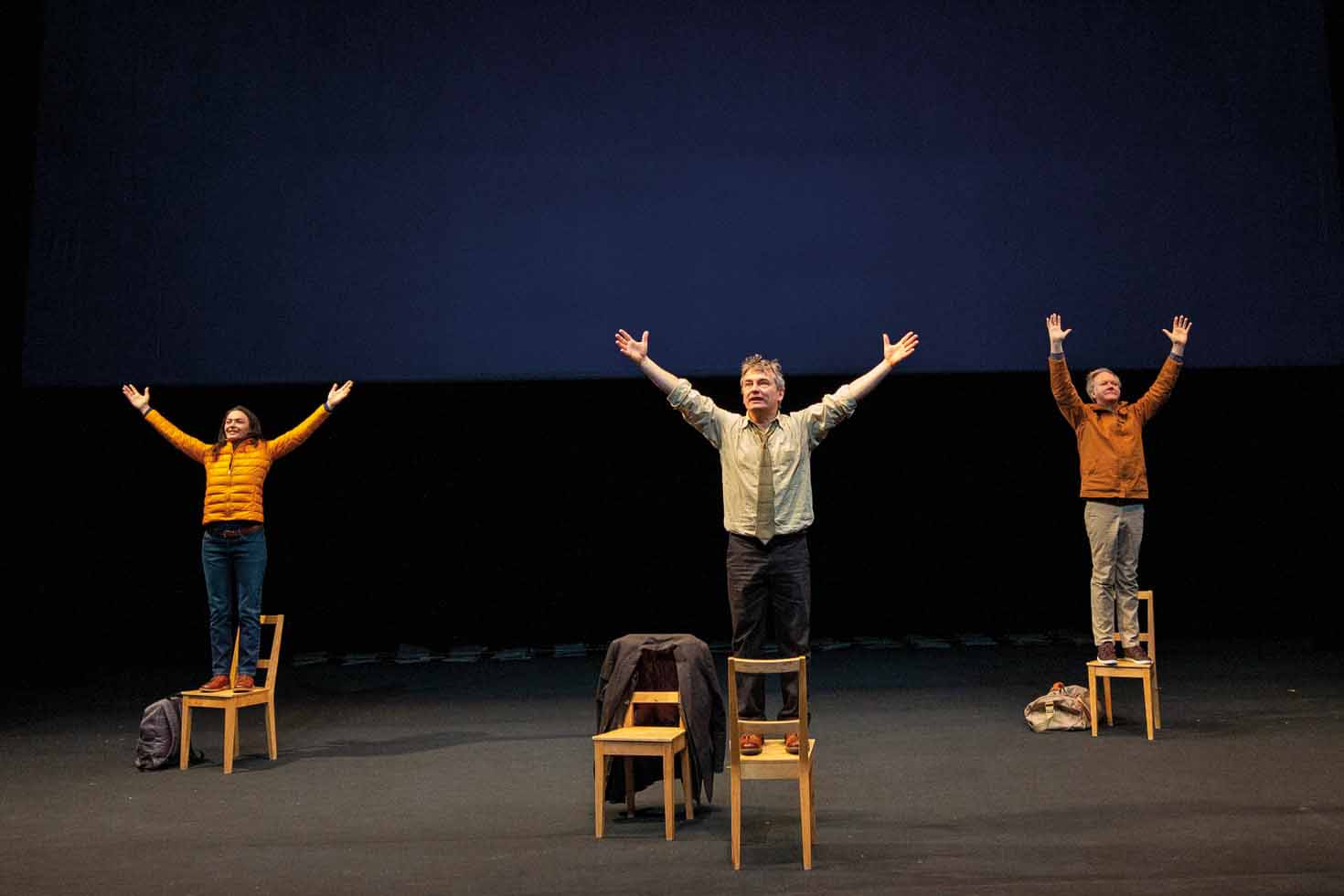 Une comédienne et deux comédiens sont chacun debout sur une chaise avec les bras en l'air.