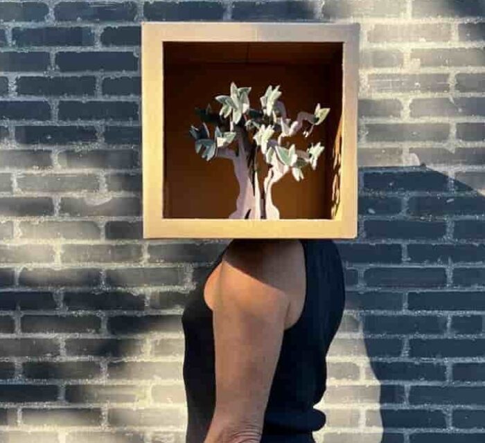 Un homme de profil devant un mur de briques noires. A la place de sa tête une boîte dont le côté est ouvert et dans laquelle on voit un arbre en 3 papier en 3D.