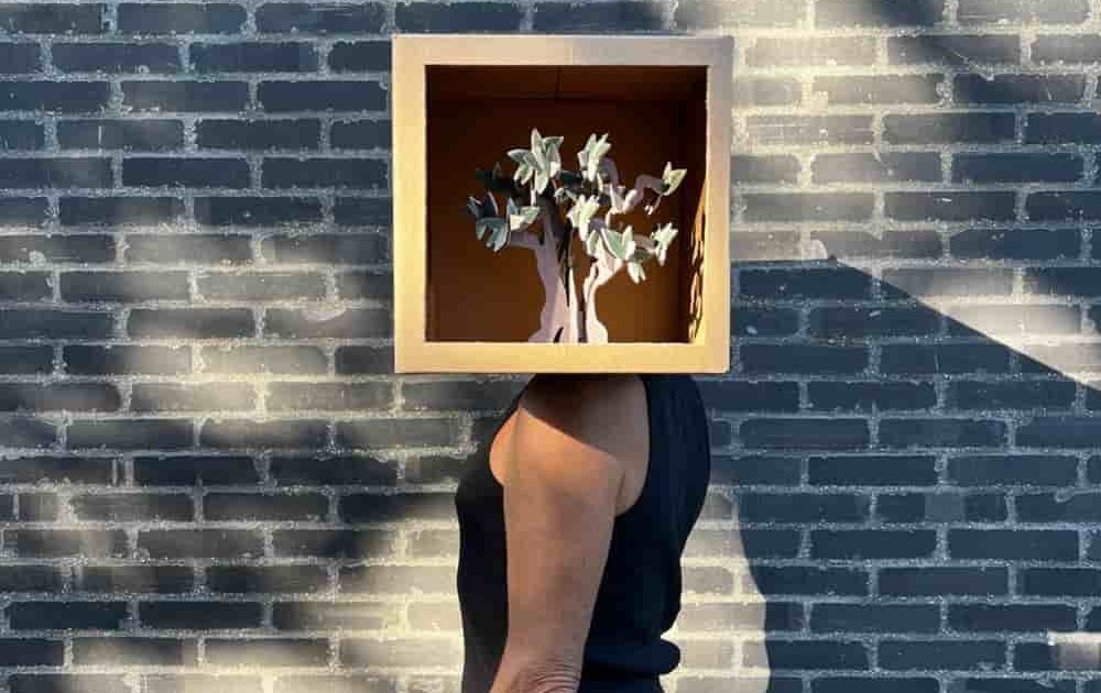 Un homme de profil devant un mur de briques noires. A la place de sa tête une boîte dont le côté est ouvert et dans laquelle on voit un arbre en 3 papier en 3D.