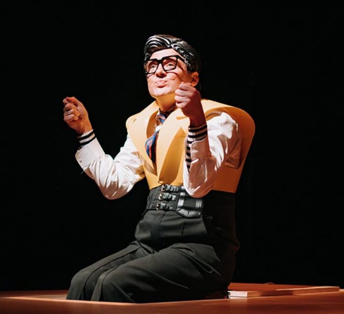 Un comédien assis, les pieds dans une trappe, porte un masque et un costume noir et jaune très marque pour incarner le personnage du père.