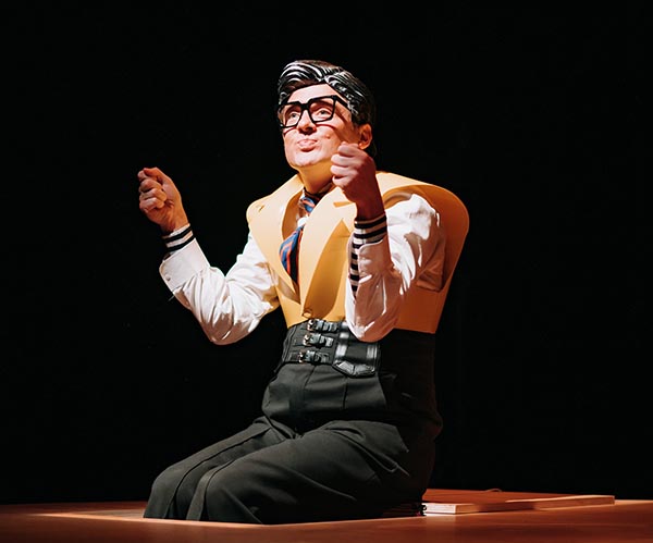 Un comédien assis, les pieds dans une trappe, porte un masque et un costume noir et jaune très marque pour incarner le personnage du père.