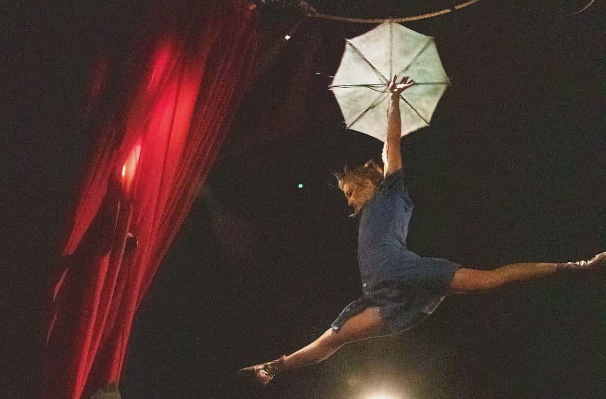 Une fil de fériste saute en grand écart avec une ombrelle à la main.