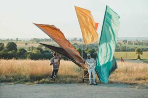 Photos du spectacle en extérieur avec le musicien jouant de la guitare électrique entouré de 4 étendards de couleur différente : noir, orangé, jaune, vert. Le comédien est visible de dos à gauche du musicien.