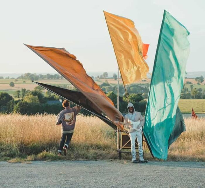 Photos du spectacle en extérieur avec le musicien jouant de la guitare électrique entouré de 4 étendards de couleur différente : noir, orangé, jaune, vert. Le comédien est visible de dos à gauche du musicien.