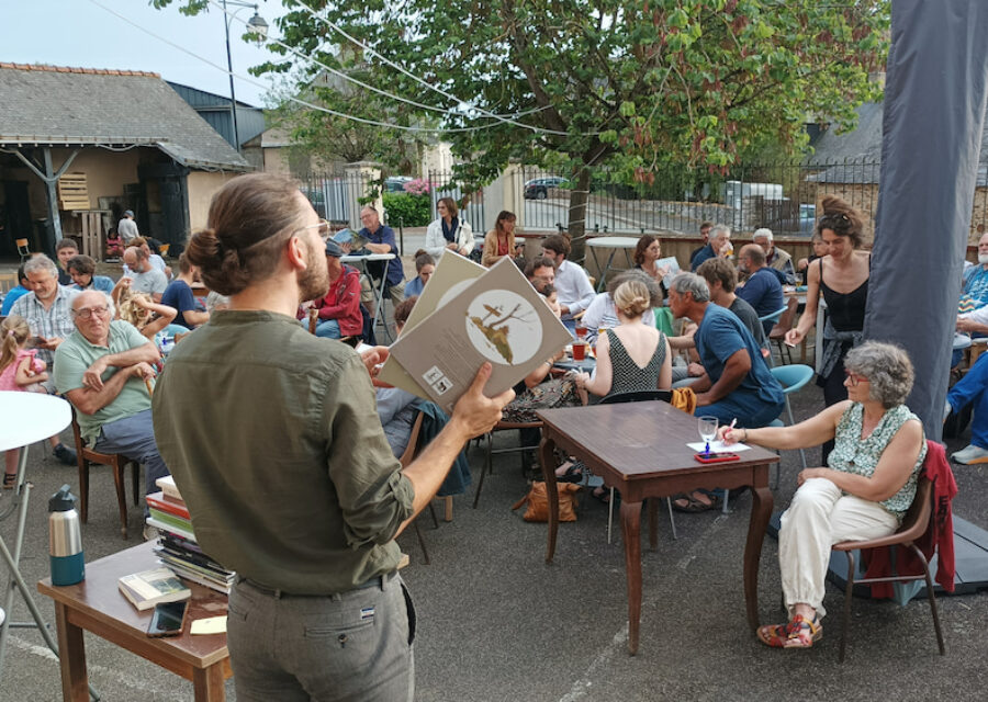 François Vincent présente des bandes-dessinées face au public installé autour de tables , dans une cour, en extérieur.