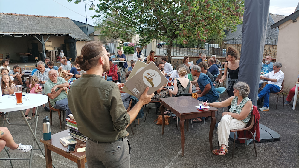 François Vincent présente des bandes-dessinées face au public installé autour de tables , dans une cour, en extérieur.