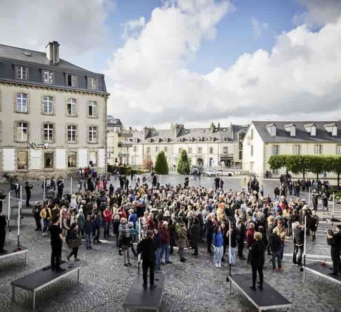 Les 20 sonneurs installés en rond sur une place avec le public debout au milieu.