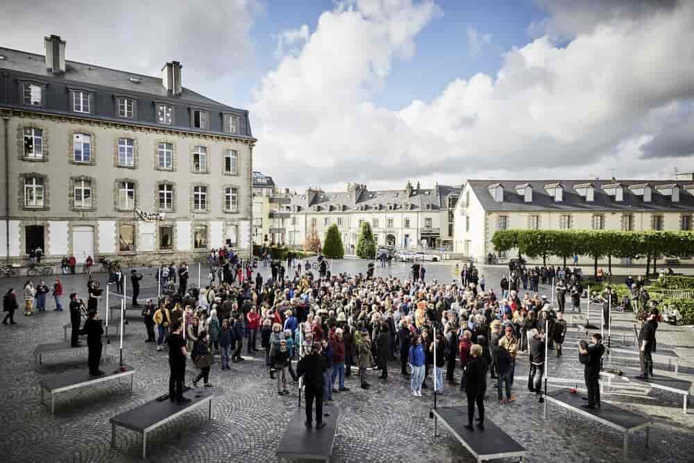 Les 20 sonneurs installés en rond sur une place avec le public debout au milieu.