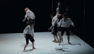 5 danseurs en bermuda noir et blouse blanche transparente ainsi qu'un musicien avec un gong en fond de scène. Deux des interprètes sont en l'air, en extension.