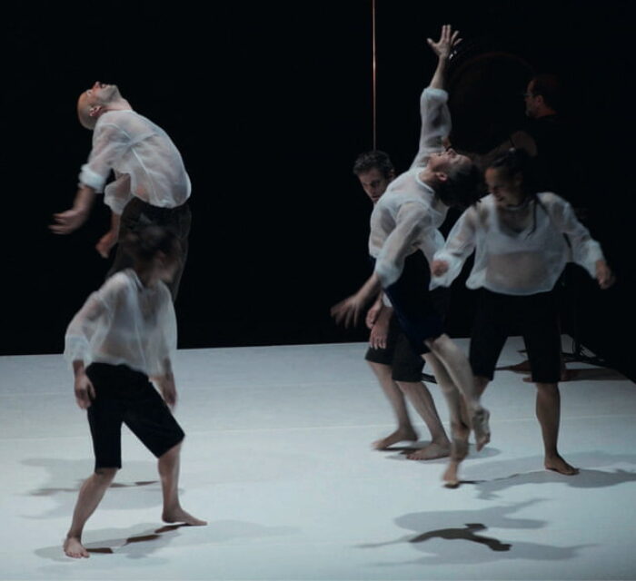 5 danseurs en bermuda noir et blouse blanche transparente ainsi qu'un musicien avec un gong en fond de scène. Deux des interprètes sont en l'air, en extension.