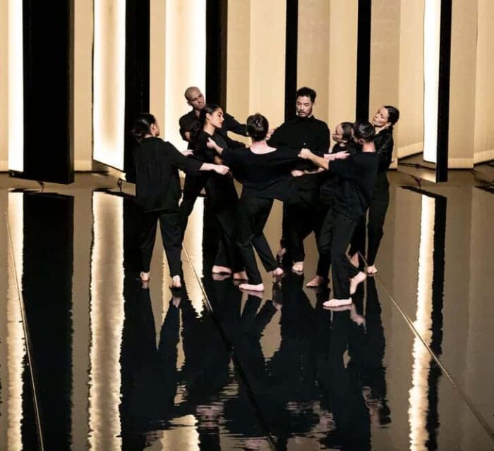 Groupe de 8 danseurs avec des pantalons et chemises noires, dans un décor contrasté entre noir et lumière.