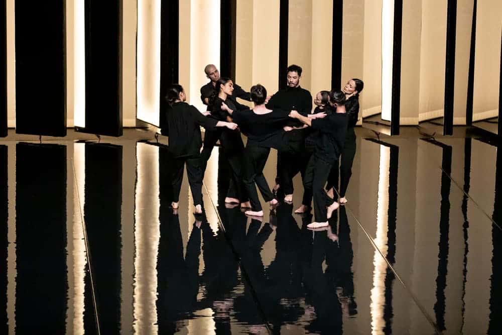 Groupe de 8 danseurs avec des pantalons et chemises noires, dans un décor contrasté entre noir et lumière.