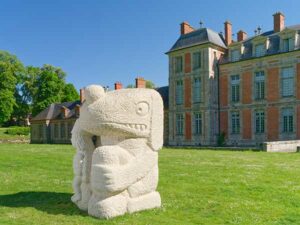 Sculpture intitulée Fate's fool, en grès de taille imposante sur la pelouse devant un château. L'oeuvre représente un personnage plutôt animal assis qui tient dans sa main droite un autre personnage humain plus petit.