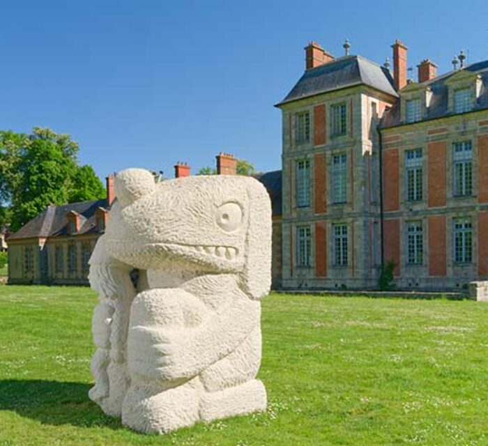Sculpture intitulée Fate's fool, en grès de taille imposante sur la pelouse devant un château. L'oeuvre représente un personnage plutôt animal assis qui tient dans sa main droite un autre personnage humain plus petit.
