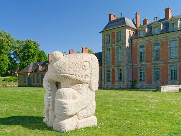 Sculpture intitulée Fate's fool, en grès de taille imposante sur la pelouse devant un château. L'oeuvre représente un personnage plutôt animal assis qui tient dans sa main droite un autre personnage humain plus petit.