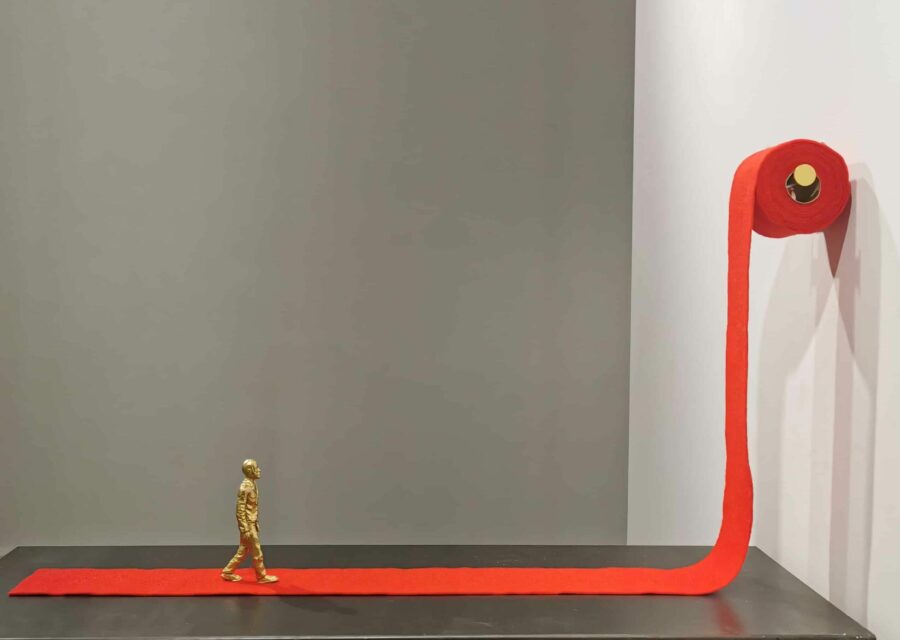 Oeuvre intitulée La Rançon de la gloire. Un sculpture composée d'un homme doré qui avance sur un tapis rouge qui provient en fait d'un rouleau de papier toilette accroché plus haut..