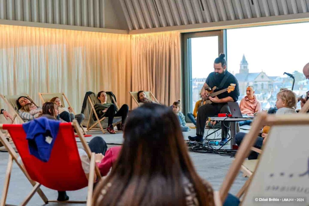 Le public est installé en cercle , assis dans des transats et on voit un musicien qui joue de la guitare électrique.