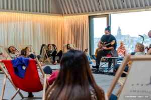 Le public est installé en cercle , assis dans des transats et on voit un musicien qui joue de la guitare électrique.