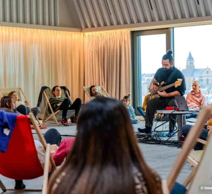 Le public est installé en cercle , assis dans des transats et on voit un musicien qui joue de la guitare électrique.